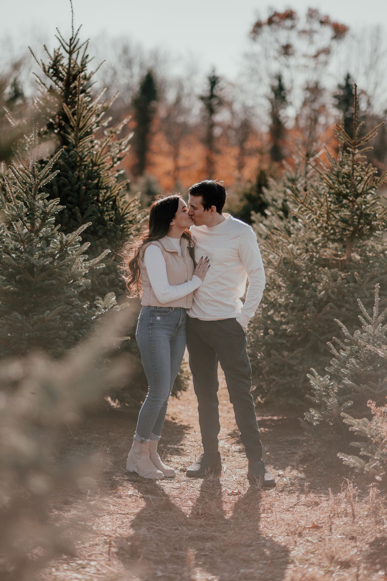 Romantic couple in casual wear embracing between evergreen trees during sunset photoshoot.
