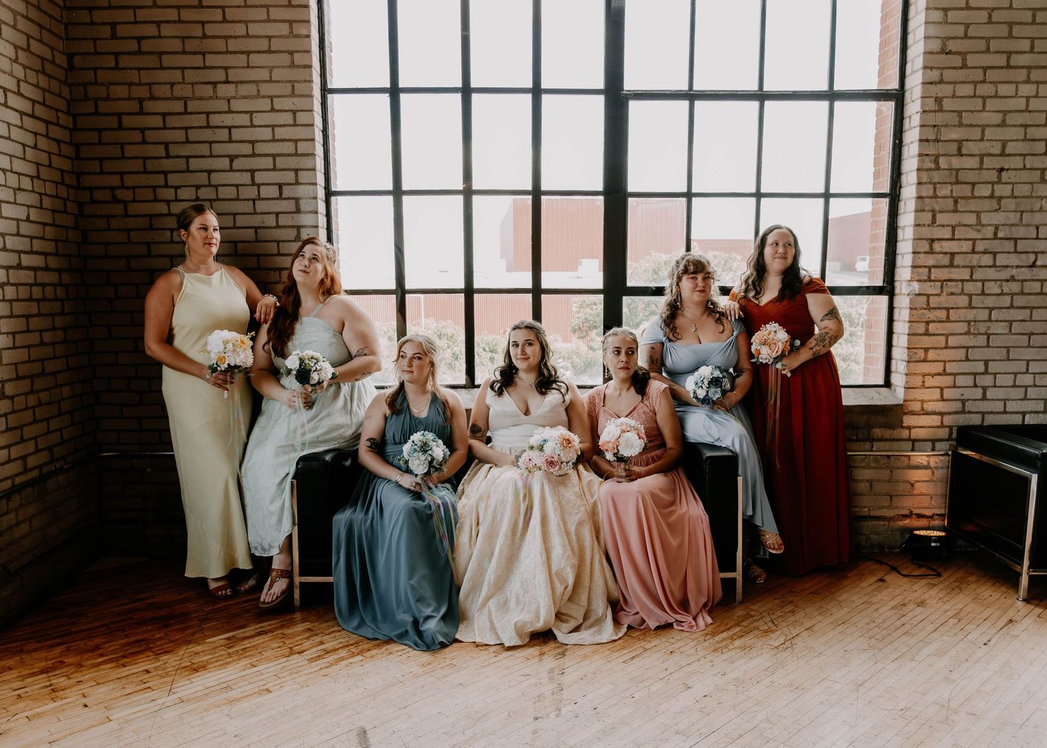 Wedding party posed in elegant dresses in varying colors beside large industrial windows in rustic brick venue.