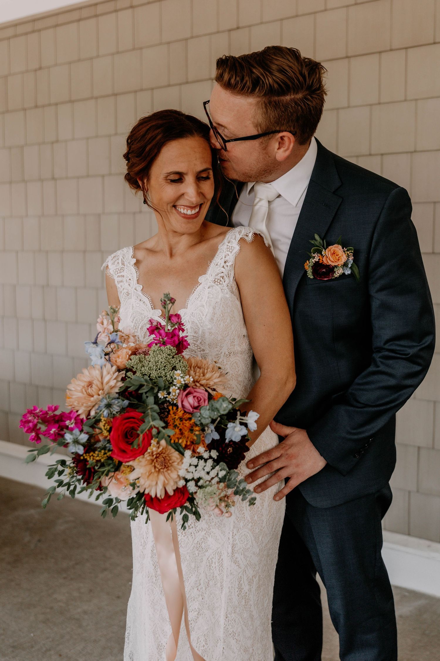 Wedding couple sharing intimate moment with colorful floral bouquet against neutral building backdrop.