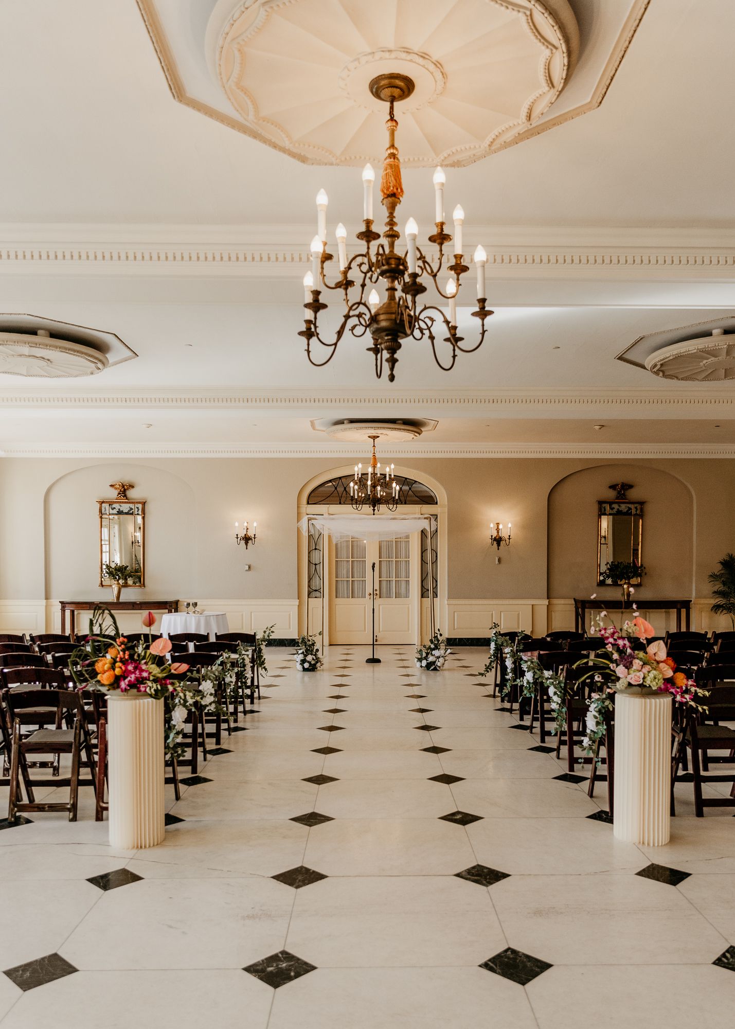 An elegant wedding ceremony space with checkered floors, chandeliers, and rows of chairs decorated with floral arrangements.