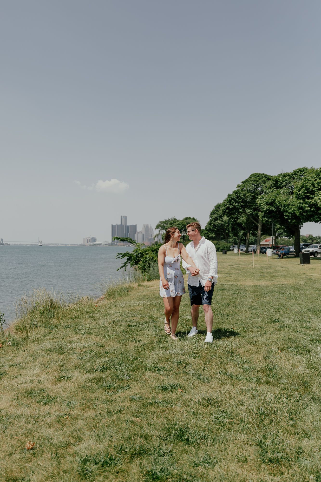 A couple standing together on a grassy waterfront with city skyline in background.