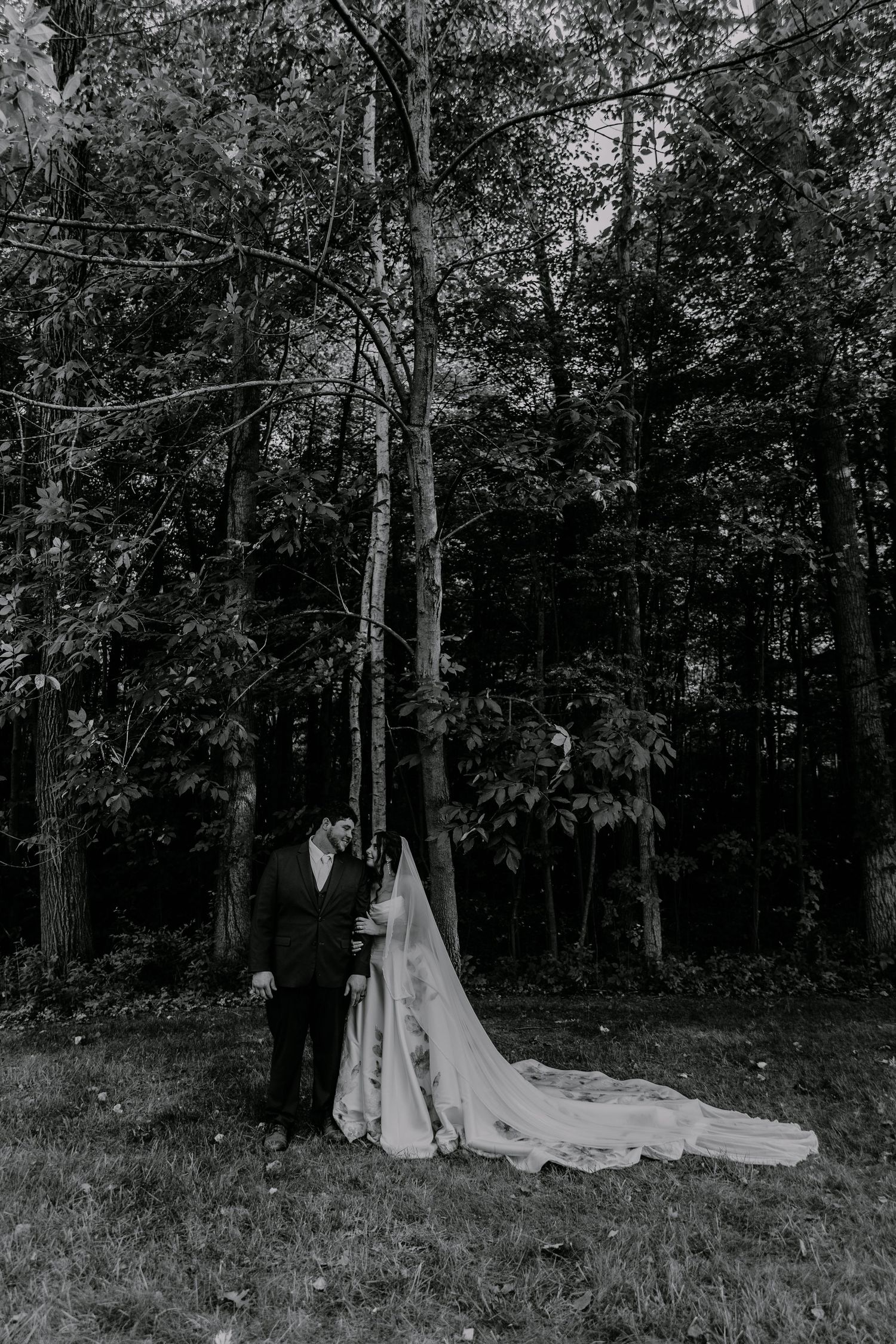 Black and white photo of bride and groom embracing by tall trees in a lush forest, bride's long veil trailing on grass.