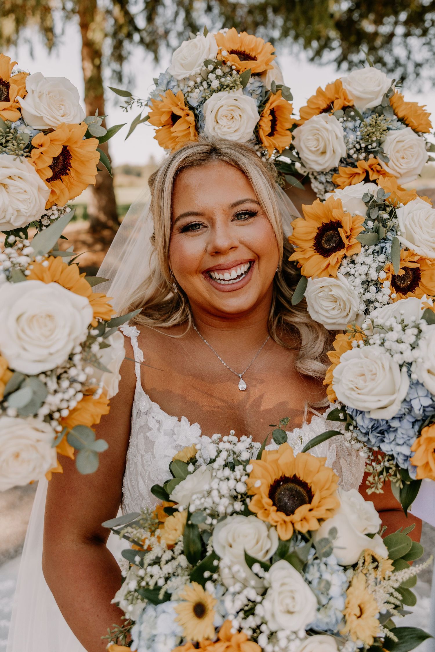 Smiling bride holding sunflower and white rose bouquet, framed by floral wreath of sunflowers and roses.