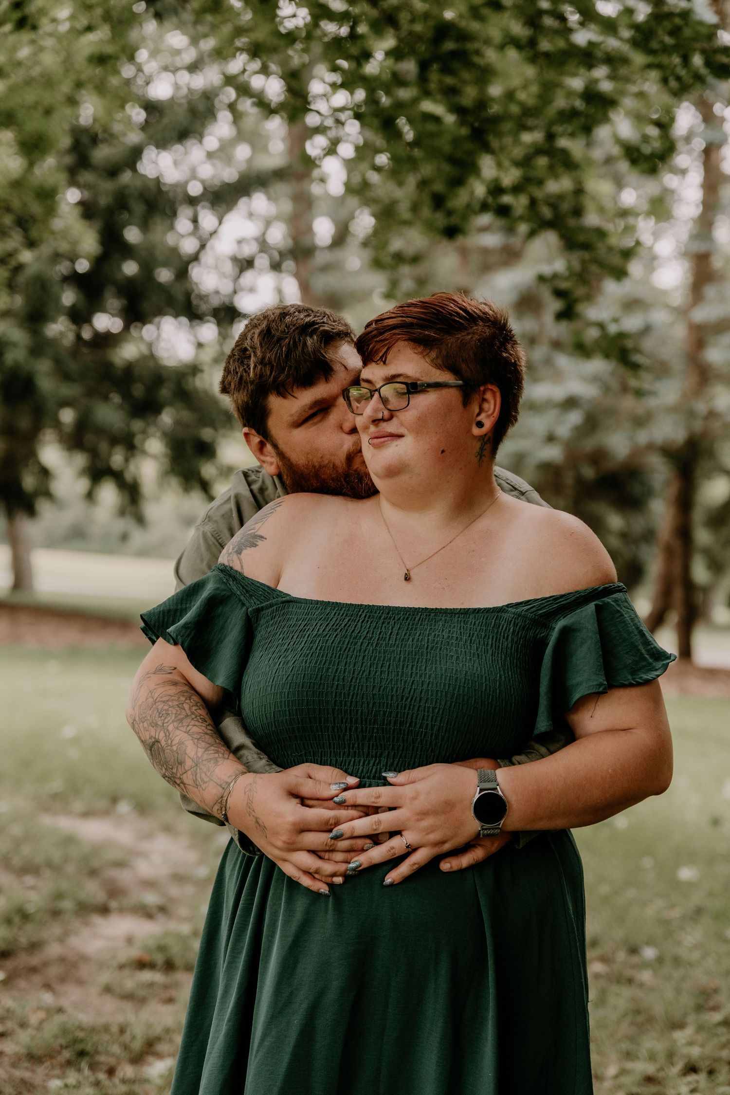 A couple embraces outdoors, man kissing woman's cheek, she wears a green off-shoulder dress, surrounded by lush trees.