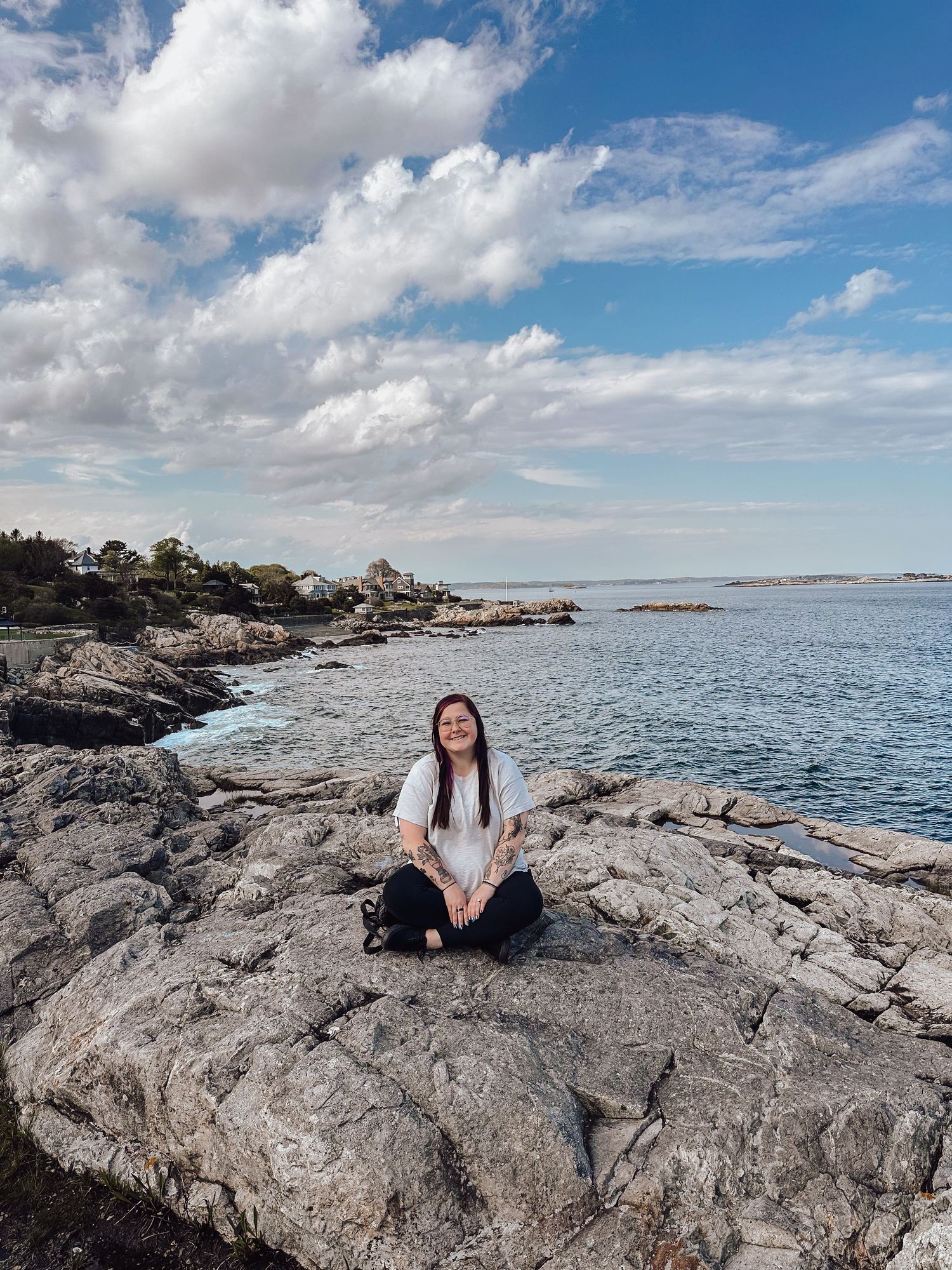 Woman sitting cross-legged on rocky coastal shore, smiling up at blue sky with white clouds and calm sea behind her.