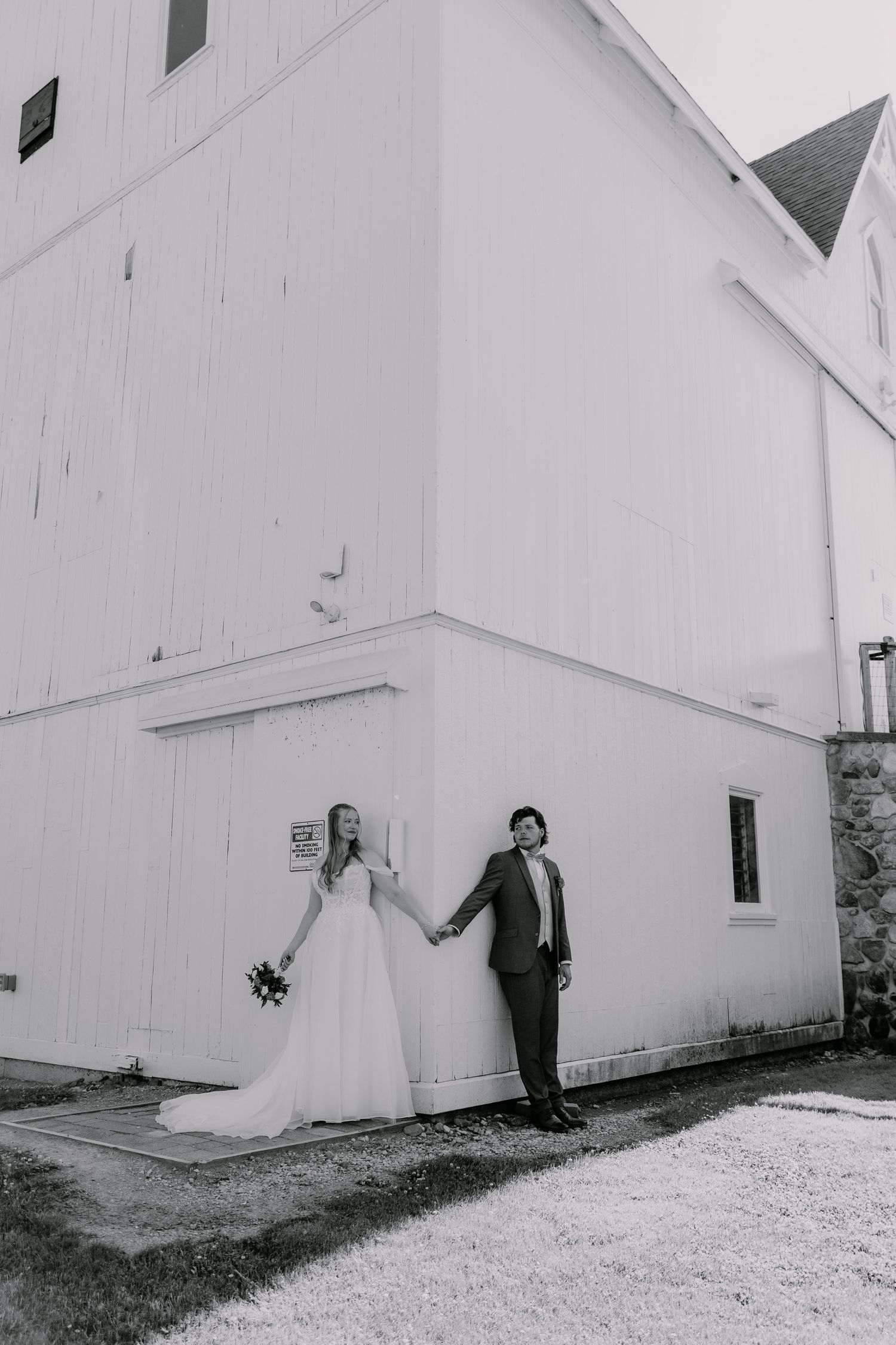 Black and white photo of bride and groom holding hands against a white barn wall on their wedding day.