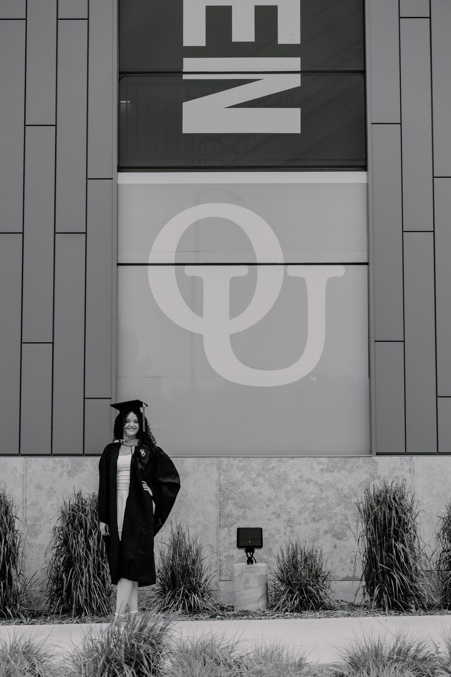 A figure in a dark coat poses against a gray building wall with 'NE' and 'QU' typography signage.