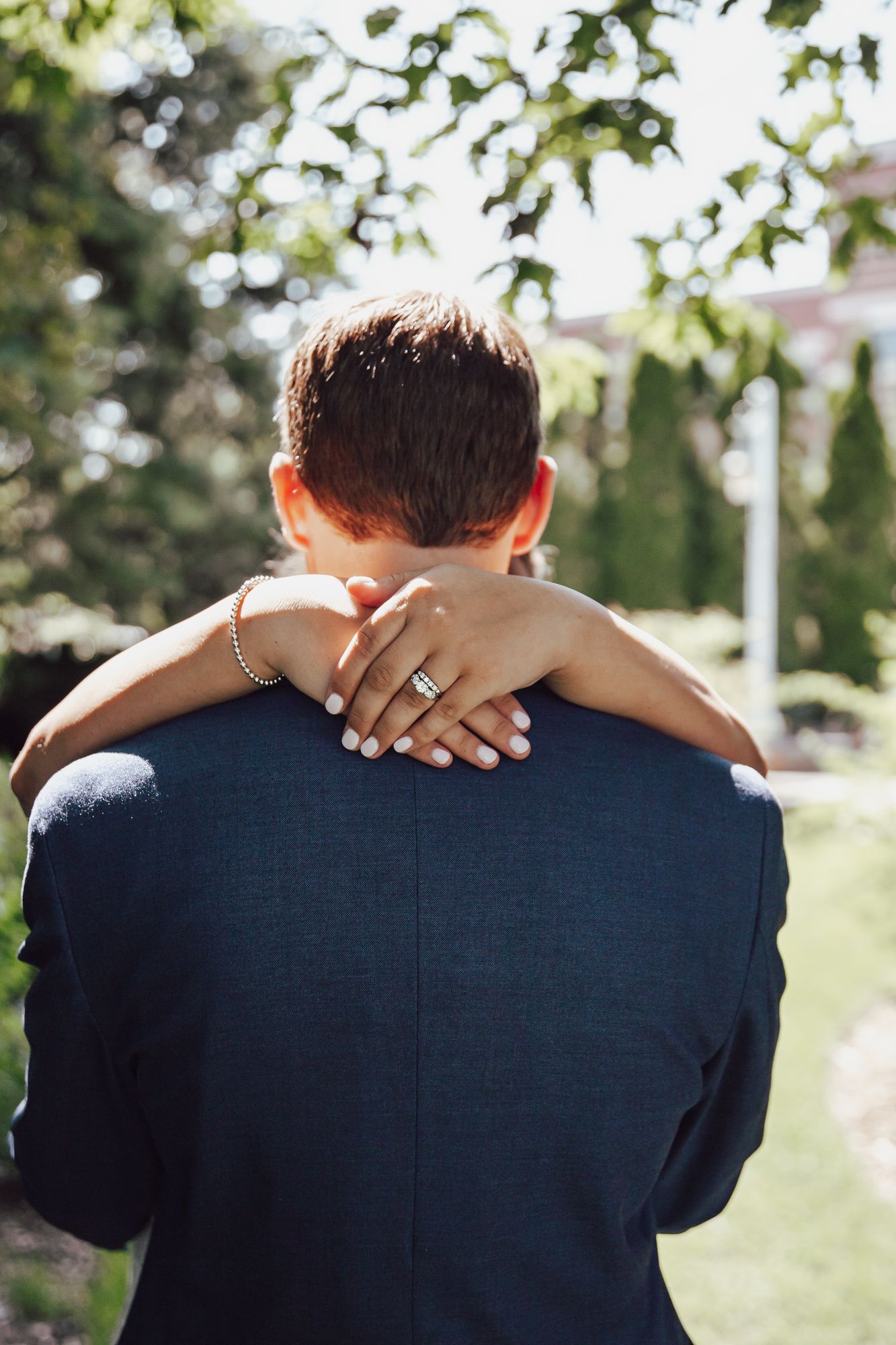 Close-up of bride's hands with wedding ring embracing groom from behind in garden setting.