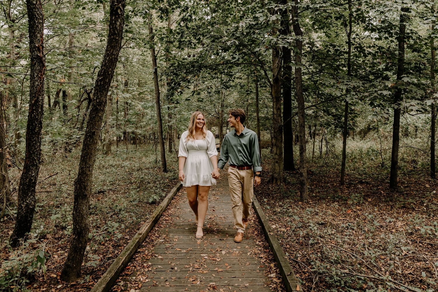 Couple walking hand in hand on railroad tracks through wooded area in engagement photos.