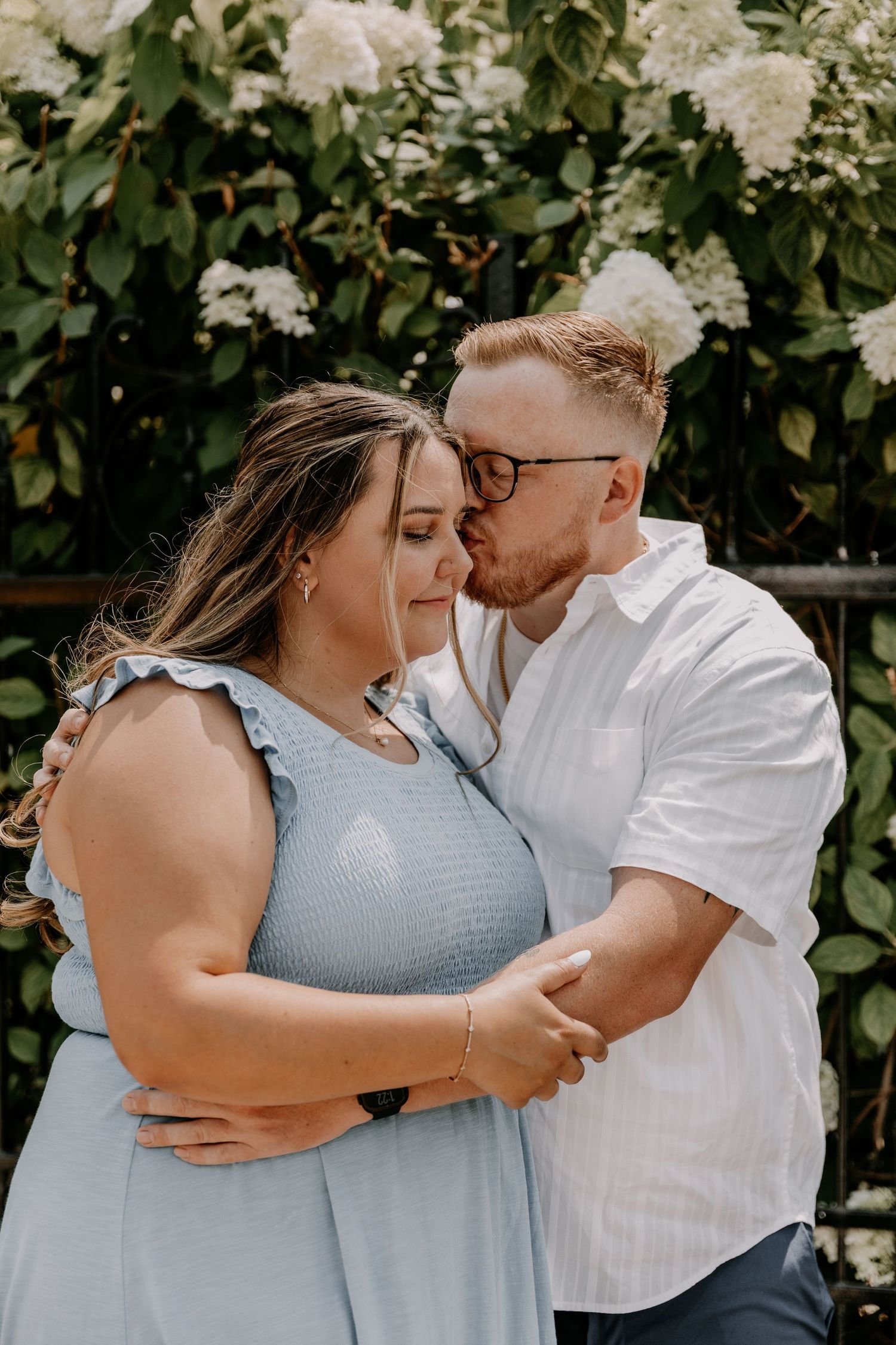 Romantic couple embracing in front of blooming white flowers, dressed in light blue dress and white shirt.