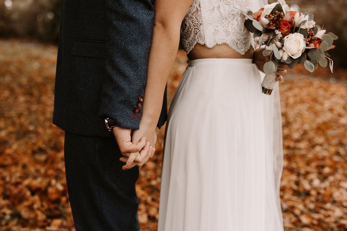 A romantic autumn wedding moment showing clasped hands and a bridal bouquet against fallen orange leaves.