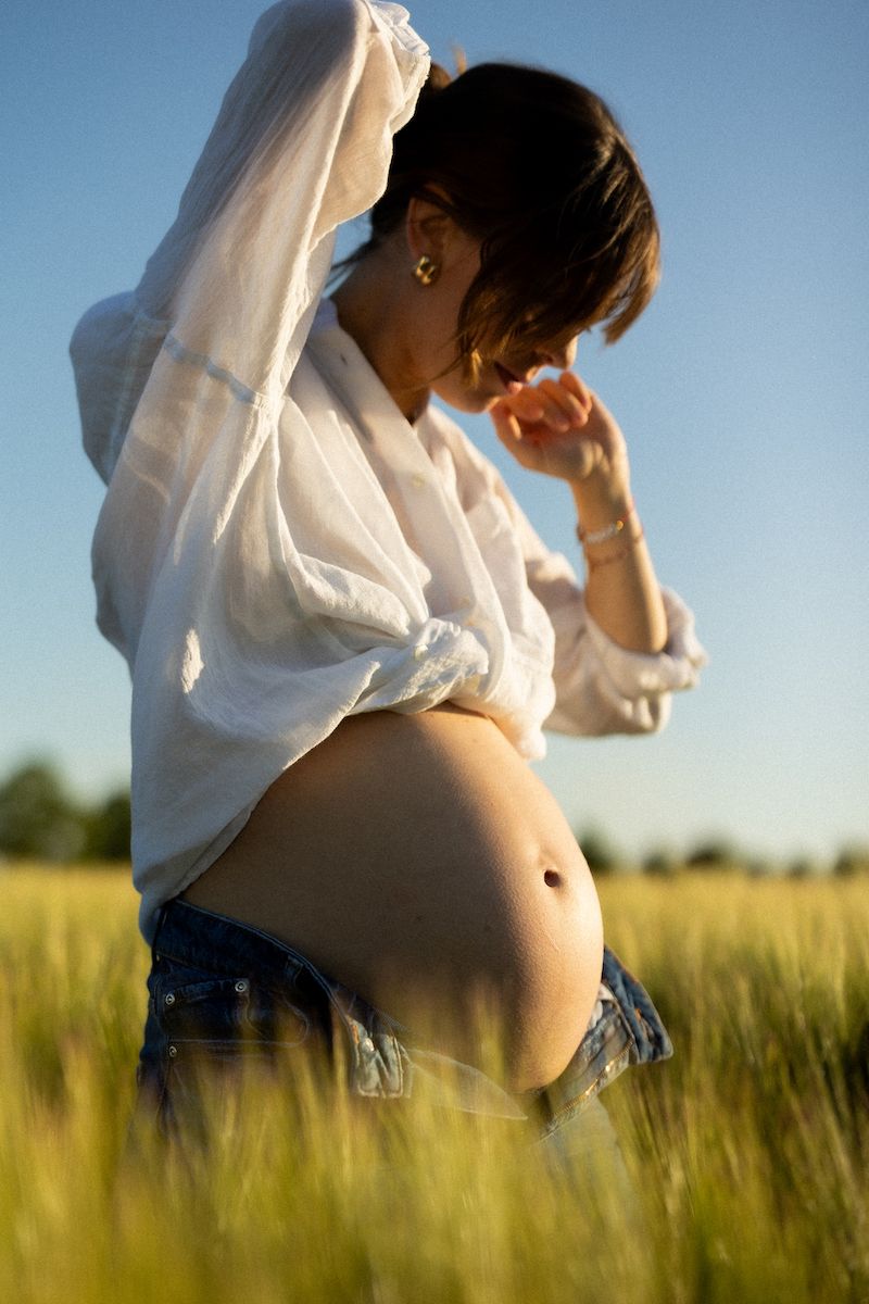 Silhouette of pregnant belly in flowing white shirt against golden field at sunset.