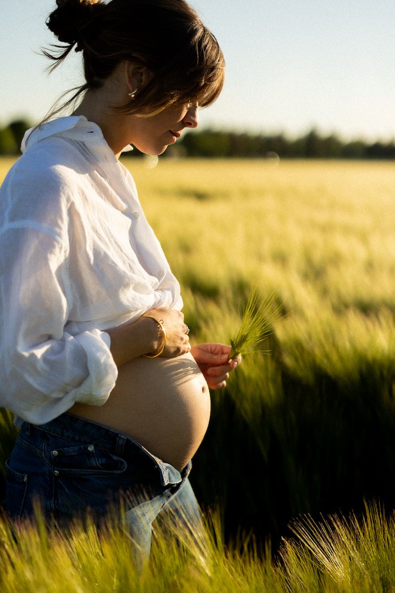 Expecting mother in white blouse and jeans stands in golden wheat field at sunset.