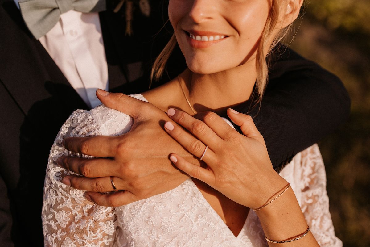 Close-up of hands with wedding rings and lace dress details in warm golden light.