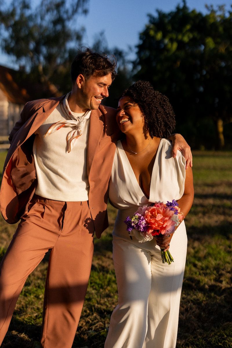 A couple laughs together in golden hour sunlight wearing coordinated white and pink formal outfits.
