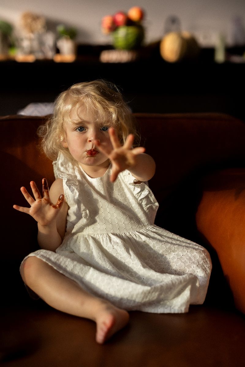 Child in white dress sitting on brown leather couch with hands raised in playful gesture.