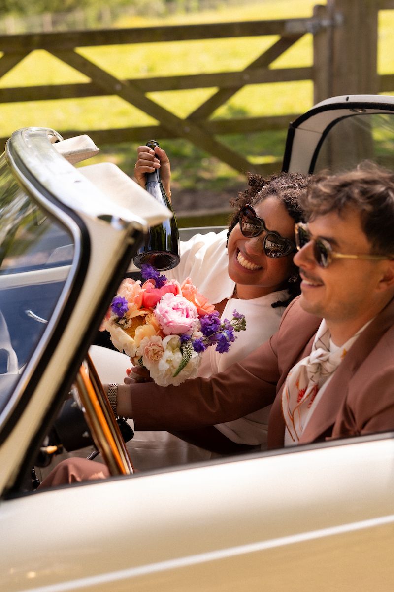 Wedding party members smile and laugh while seated in a vintage car with colorful bouquets.