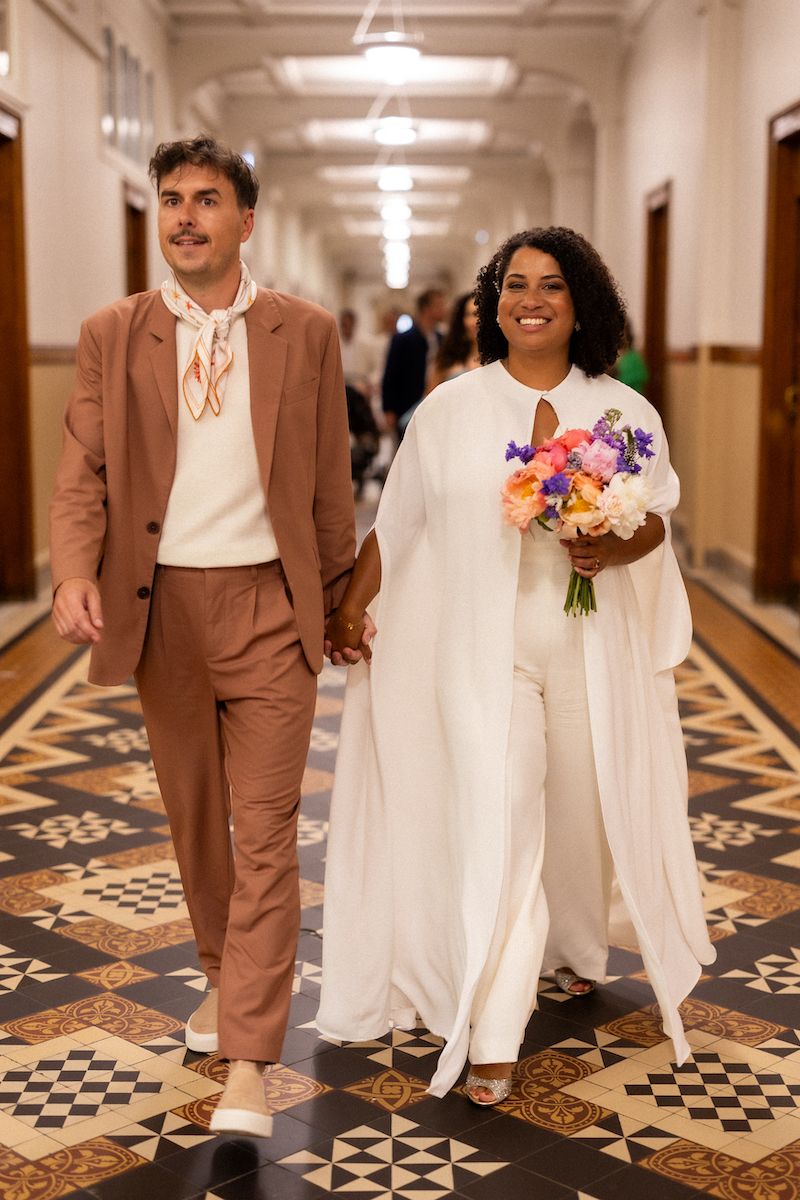 A couple walks down a hallway with decorative tiled floors, wearing white and dusty pink formal attire and holding flowers.