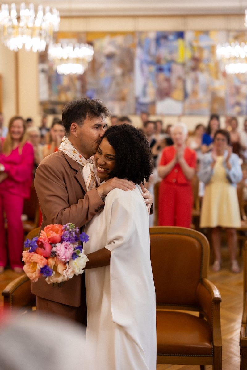 Newlyweds share intimate moment at wedding reception with colorful bouquet.
