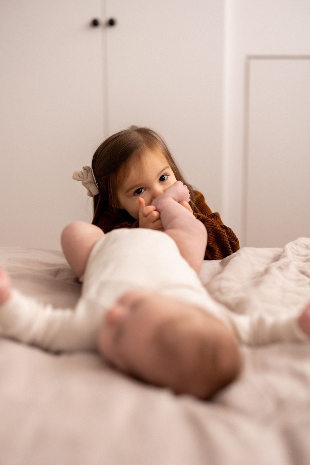 A child in a brown sweater lying on a bed next to a baby.