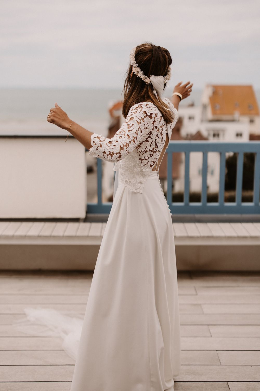 Person in flowing white lace dress twirling on waterfront deck with buildings in background.