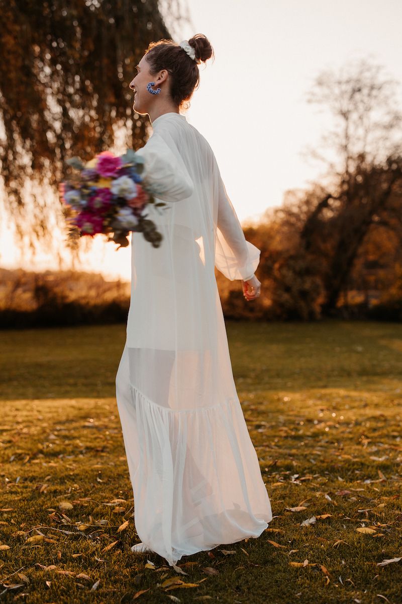 Bride in flowing white dress holding colorful bouquet during golden hour.