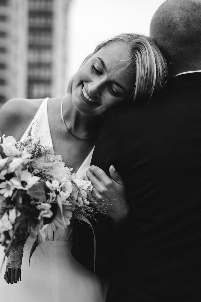 A candid black and white moment of tenderness between newlyweds with a bouquet.