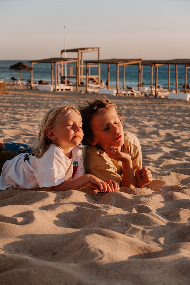 Two children lying on beach sand at sunset with wooden structures in background.