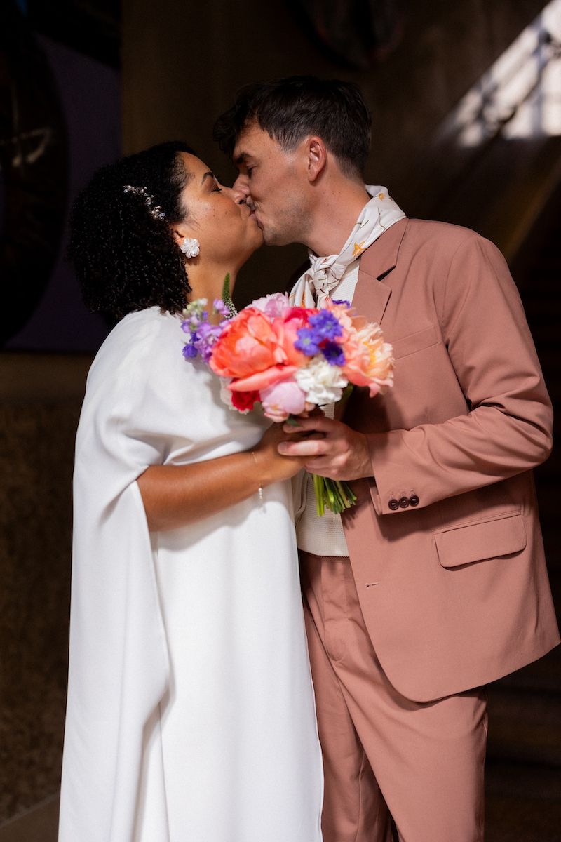 Newlyweds share a kiss in a corridor, with one in white and one in a pink suit holding a vibrant bouquet.