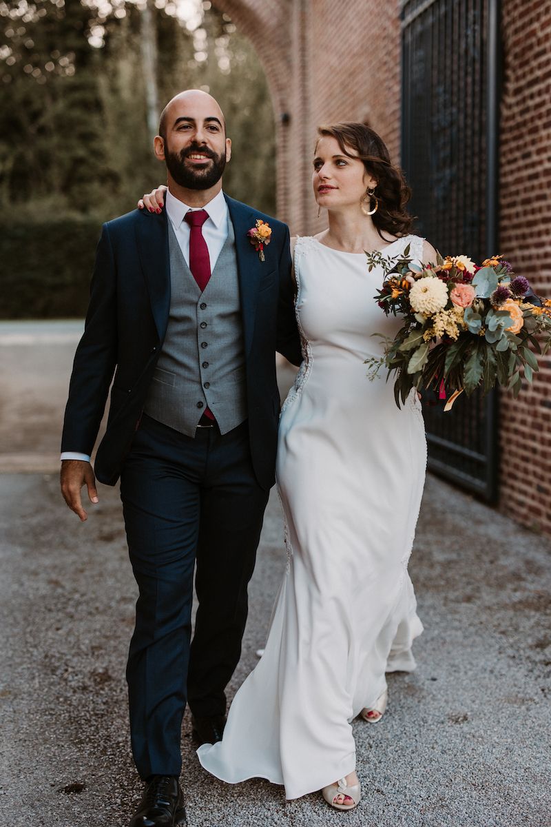 Elegant wedding couple walking together with bride holding autumn bouquet against brick building.