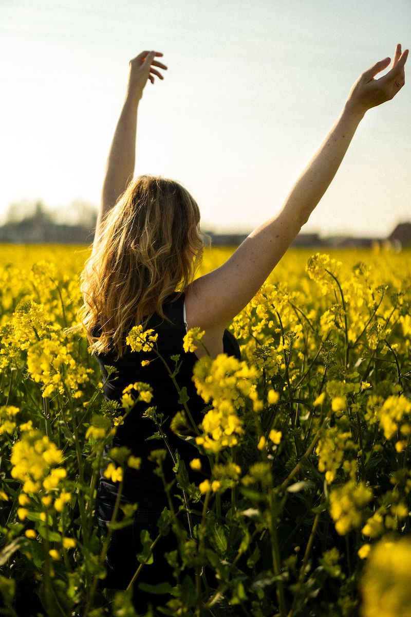 Woman with outstretched arms stands in a blooming yellow canola field at golden hour.