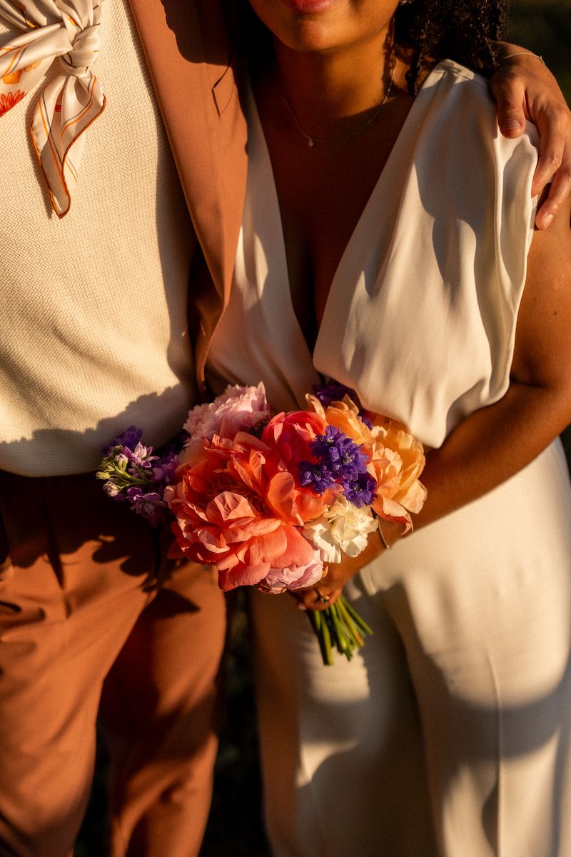 Close-up of a colorful wedding bouquet with coral and purple flowers against white and pink formal wear.