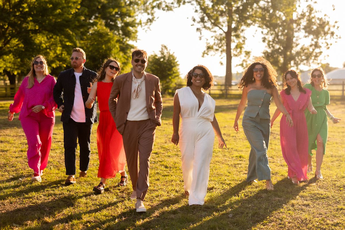 Wedding party walks together across sunlit grass in colorful dresses creating silhouettes.