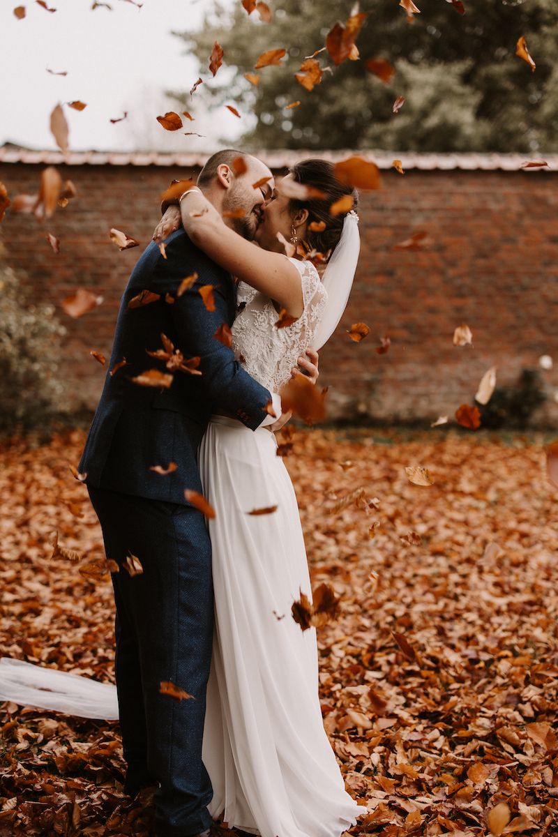 A romantic autumn wedding photo with falling leaves as couple embraces in front of brick wall.