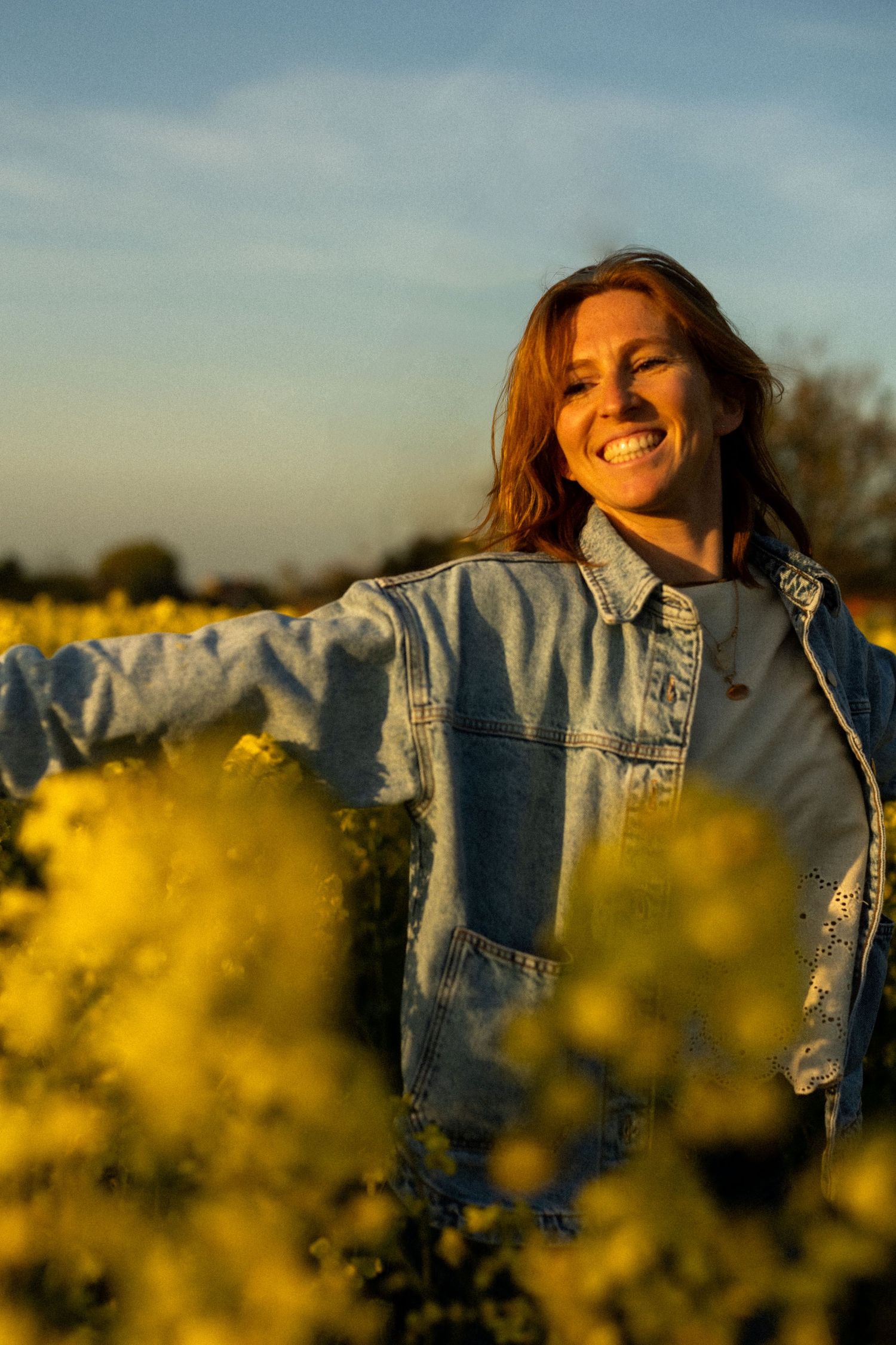 A person wearing denim smiles in a field of yellow wildflowers during golden hour.