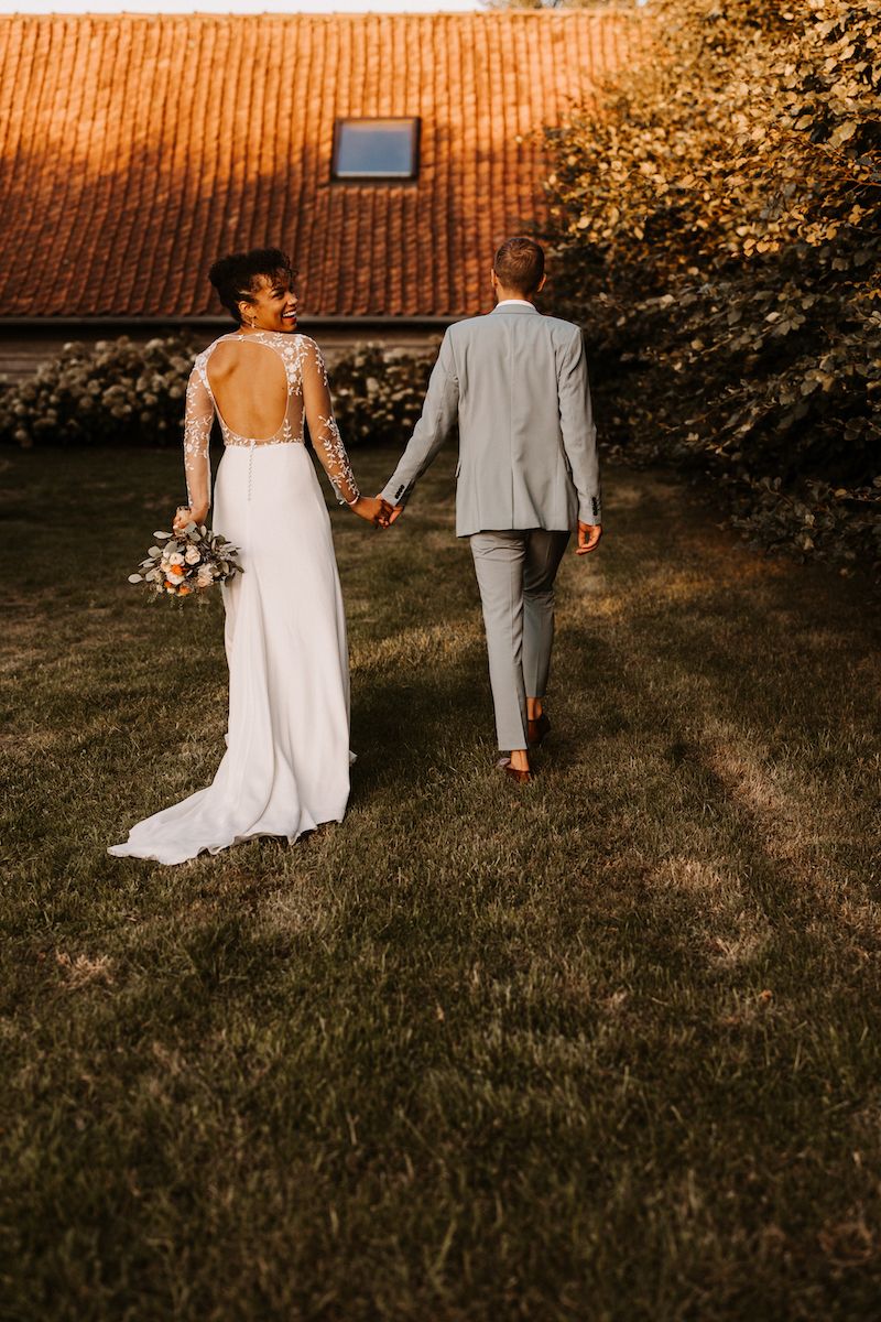 Newlyweds holding hands walking away on grass, bride in backless dress with flowing train.