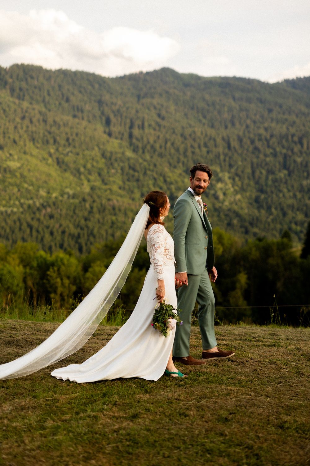 A couple in wedding attire standing on a mountainside with forest views.