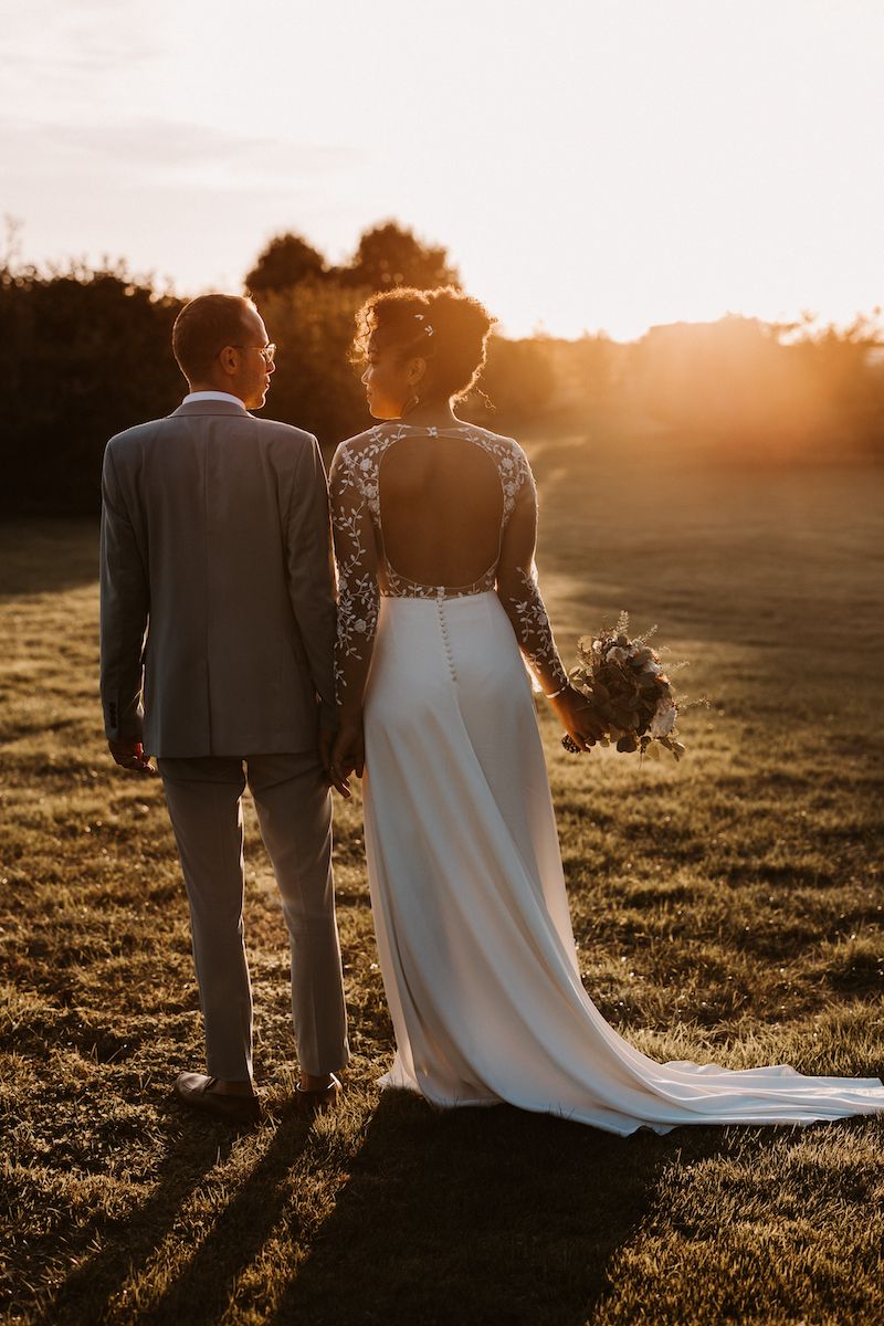 A couple in wedding attire stand together in a golden sunset field holding hands with flowing dress and suit silhouettes.