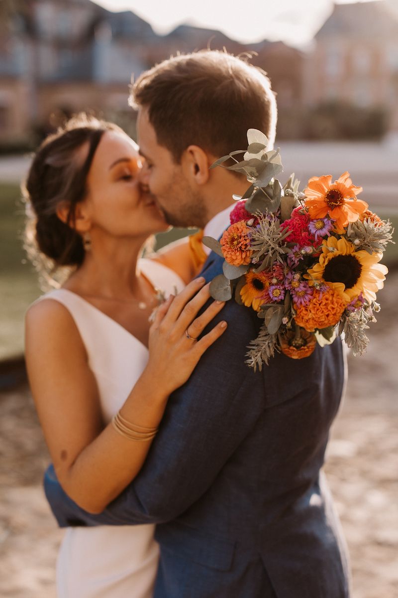 Newlyweds share a sunset kiss while holding a beautiful fall-themed bouquet with sunflowers.