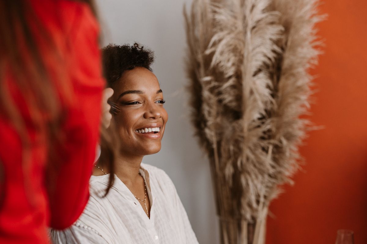 Woman in white robe smiling warmly near decorative pampas grass and orange wall.
