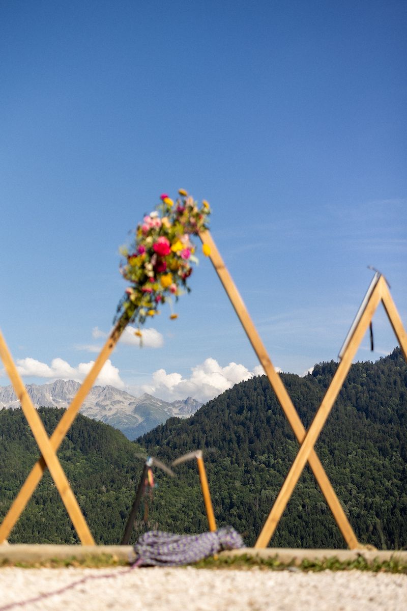Wooden wedding arch decorated with flowers against mountain backdrop in clear blue sky.
