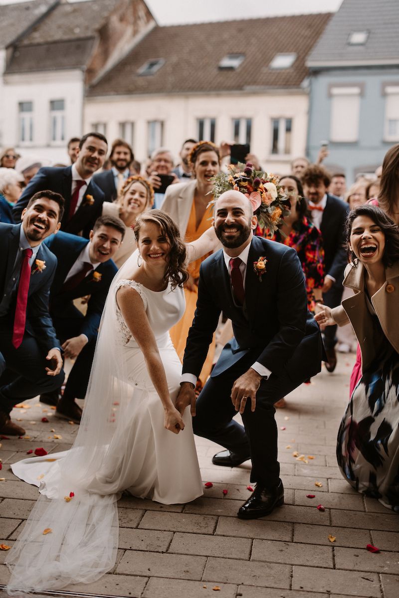 Wedding celebration with guests cheering as couple kneels on cobblestone street.