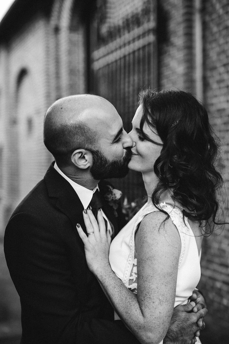 A black and white intimate wedding portrait of a couple kissing against a brick building backdrop.