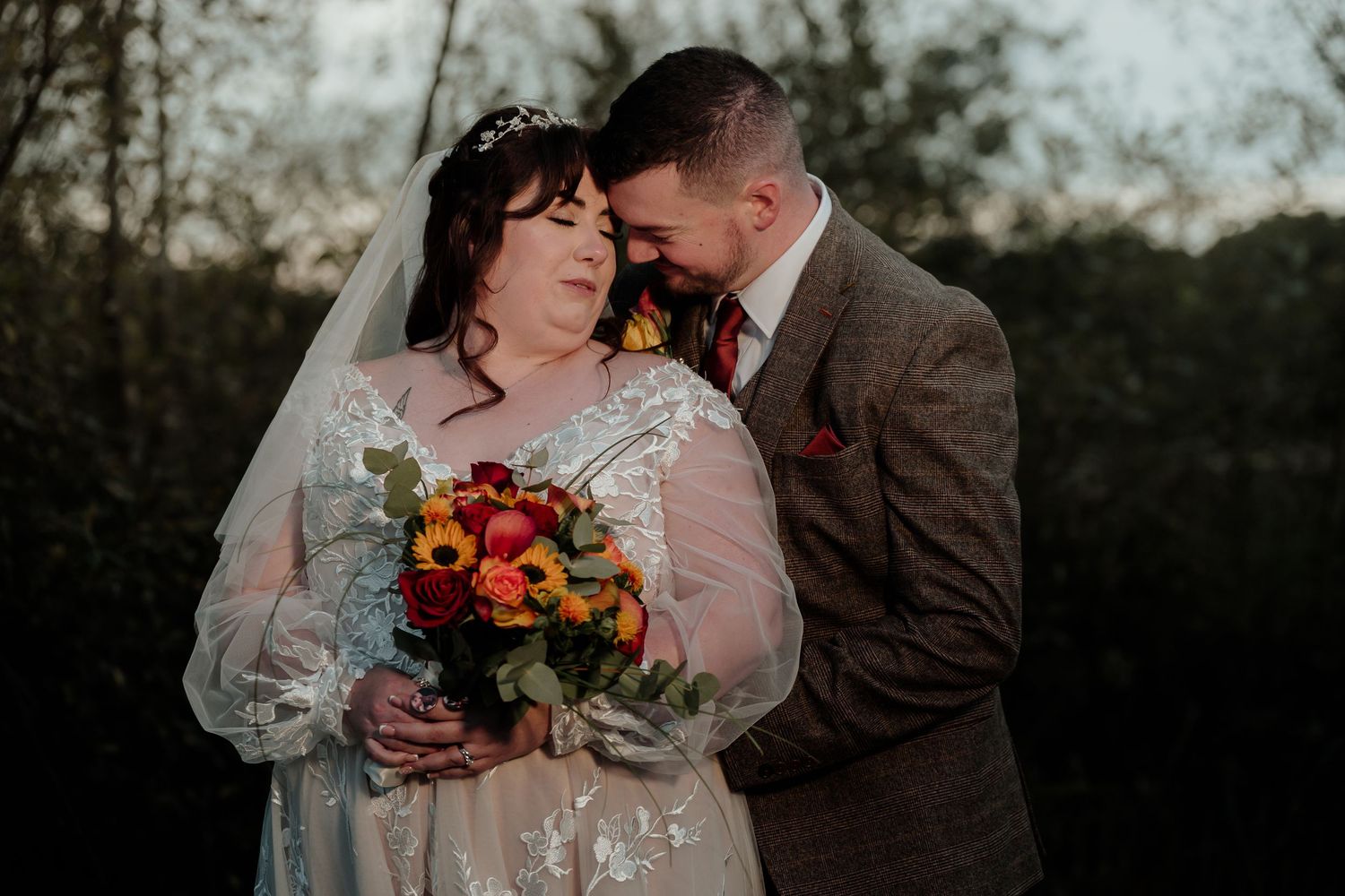 A tender autumn wedding portrait featuring a couple holding a vibrant bouquet at dusk.