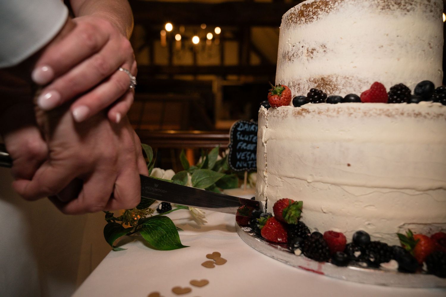 A wedding cake decorated with fresh berries and flowers being cut by newlyweds.