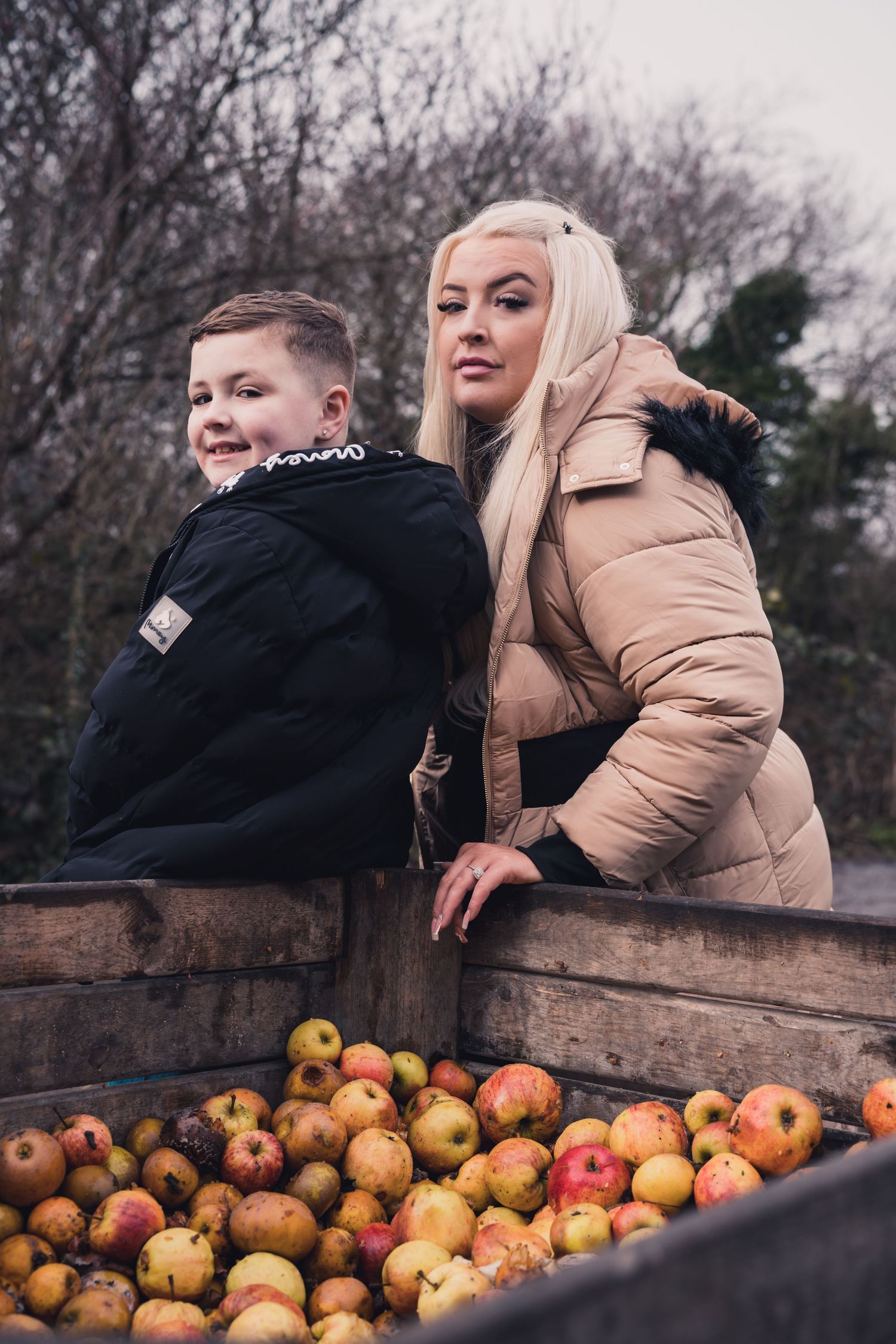 Two people posing by wooden crate filled with fresh red apples outdoors.