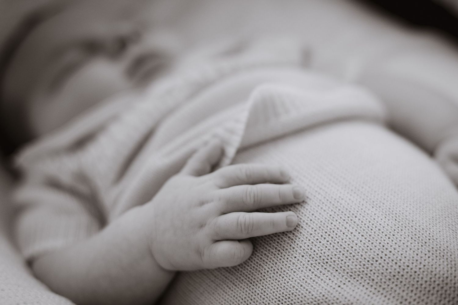 Baby's small hand rests on soft gray fabric in monochrome photograph.
