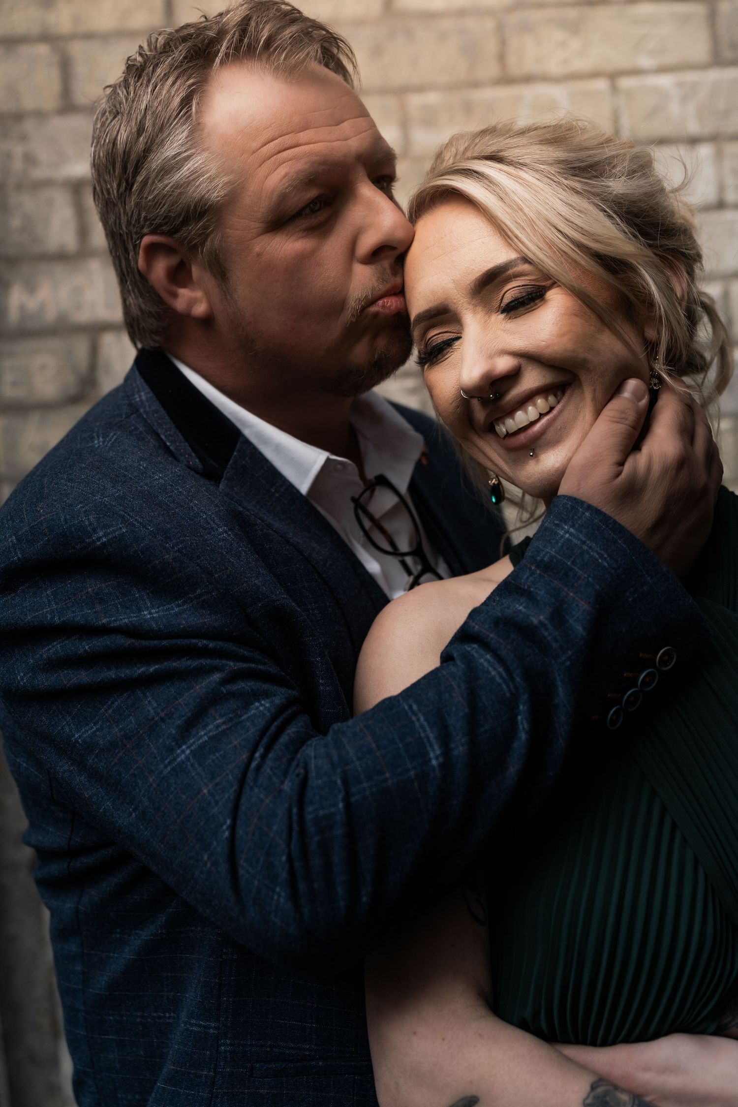 Intimate couple shares tender moment against brick wall as she smiles in green sweater while he wears navy suit.