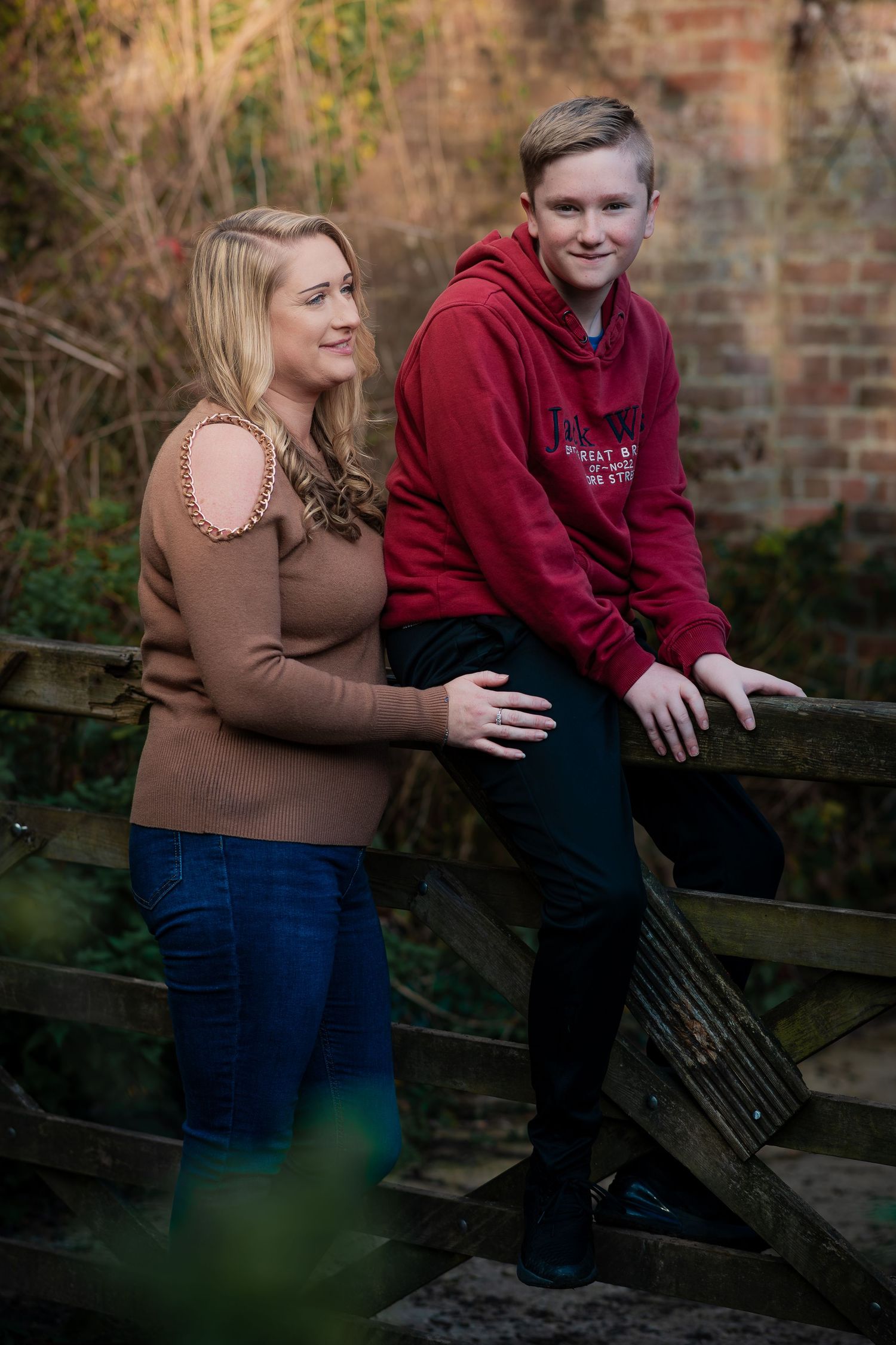 Two people in casual attire share a moment on a rustic wooden fence with brick wall backdrop in an outdoor setting.