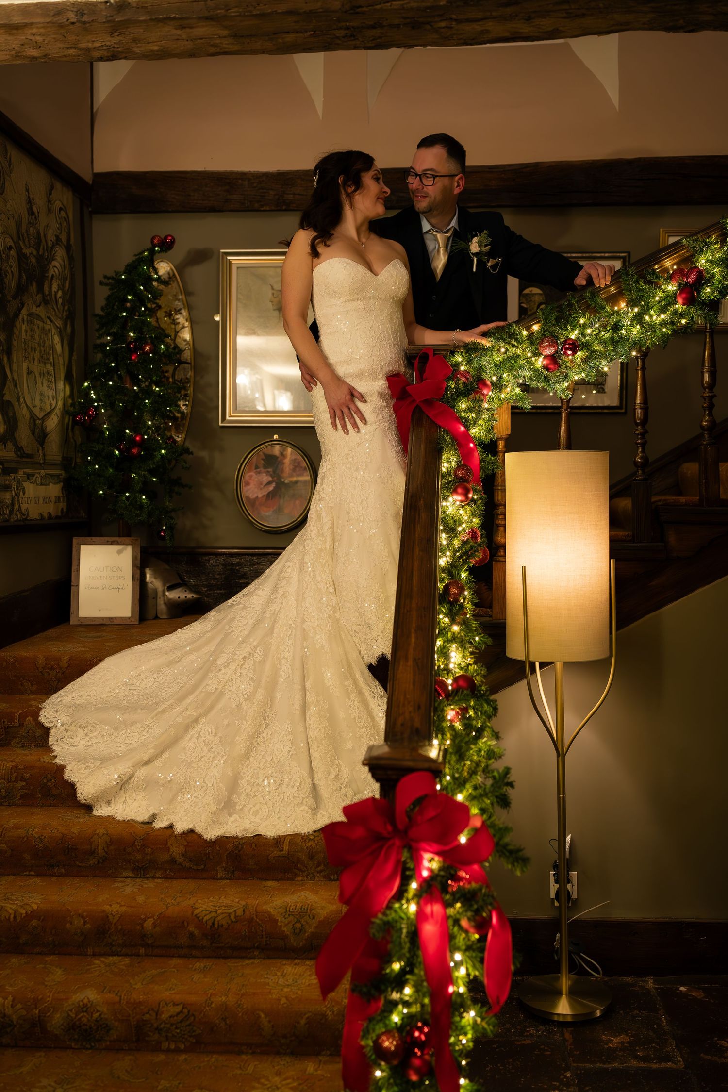 A wedding couple poses on stairs decorated with festive garlands, poinsettias and Christmas lights in a rustic venue.